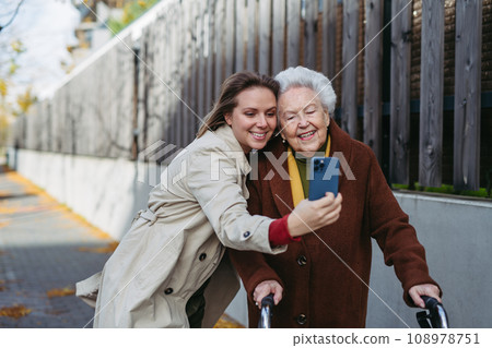 Grandmother and mature granddaughter on a walk in city streets, taking selfie. Caregiver and senior lady enjoying the windy autumn weather. 108978751