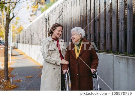Grandmother and mature granddaughter on a walk in city streets, during windy autumn day. Caregiver and senior lady with mobility walker enjoying the fall weather. 108978759
