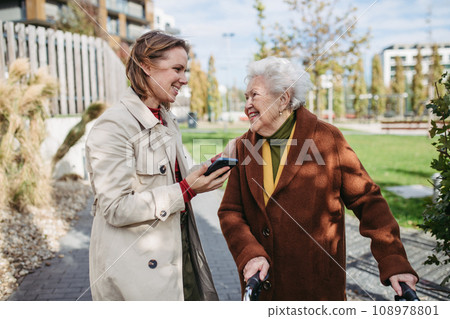 Grandmother and mature granddaughter on a walk in city park, during windy autumn day. Caregiver and senior lady with mobility walker talking and enjoying the fall weather. 108978801