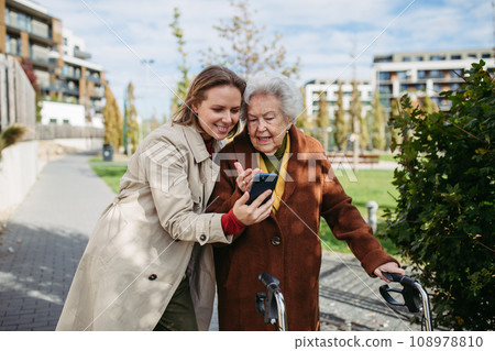 Mature granddaughter teaching grandmother how to use smartphone. Caregiver helping senior lady shopping online on smartphone. Mature granddaughter teaching grandmother how to use smartphone. Caregiver helping senior lady shopping online on smartphone. 108978810