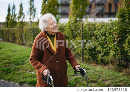 Senior woman with a mobility walker walking on city streets during autumn day, enjoying the beautiful sunny weather. Elderly lady savoring every moment, living life to fullest. 108978816