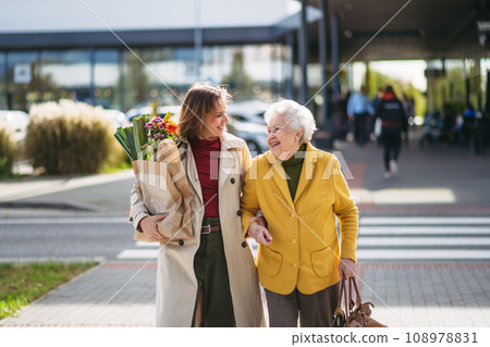 Mature granddaughter carrying groceries out to her grandmother's car. Senior woman shopping at the shopping center, needing help loading groceries into car. Mature granddaughter carrying groceries out to her grandmother's car. Senior woman shopping at the shopping center, needing help loading groceries into car. 108978831