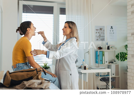 Doctor examining young patient's sore throat, looking into the throat using wooden tongue depressor. Doctor examining young patient's sore throat, looking into the throat using wooden tongue depressor. 108978977