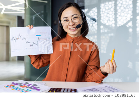 Webcam view, online video call, young asian woman at workplace inside office smiling, looking at camera, showing colleagues successful achievement results graph, businesswoman with headset phone. 108981165
