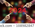 2024, People pointing to the number 2024 on background of faded lights. Happy New Year 108982847