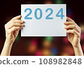 2024, Woman holding cardboard with number 2024 on background of faded lights. Happy New Year 108982848