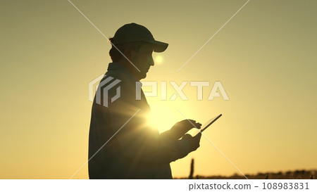 Embracing golden breathtaking sunset, modern farmer harnesses power digital technologies revolutionize agriculture. Witness silhouette dedicated agronomist work wonders field, using tablet optimize 108983831