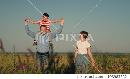 mom dad and child walking in the park. a happy family. the son sits on the shoulders of the father. children's walk with parents in a flowering park at sunset on a summer evening. teamwork. a group of 108983832