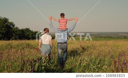 mom dad and child walking in the park. a happy family. the son sits on the shoulders of the father. children's walk with parents in a flowering park at sunset on a summer evening. teamwork. a group of 108983833