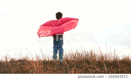 Boy stands on field straightening arms with red raincoat in cloudy weather Boy stands on field straightening arms with red raincoat in cloudy weather 108983872