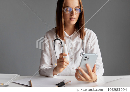 Healthcare practitioner's workplace. Professional woman doctor wearing white medical lab coat using smart phone for online consultation prescribes spray for patient telling how to take it. 108983994