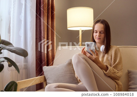 Calm brown haired young Caucasian woman using mobile phone while sitting couch at home scrolling online browsing web pages checking social network or texting with friends. Calm brown haired young Caucasian woman using mobile phone while sitting couch at home scrolling online browsing web pages checking social network or texting with friends. 108984018