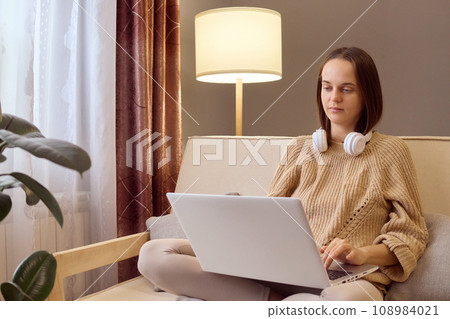 Brown haired woman in knitted sweater using laptop sitting on couch at home beautiful girl shopping or chatting online in social network watching movie freelancer working on computer project. 108984021