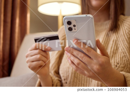 Unrecognizable woman holding smartphone and banking credit card involved in online mobile shopping at home female shopper purchasing goods or services in internet store. 108984053