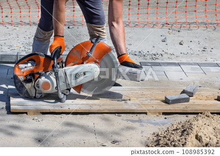 A worker cuts paving slabs with a diamond blade and a gas cutter on a clear summer day. 108985392