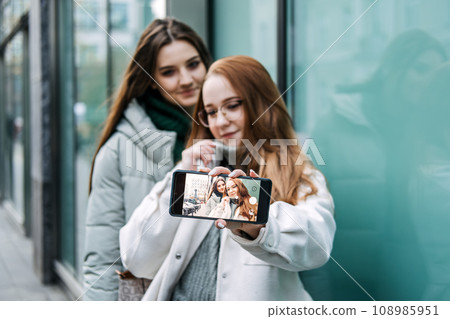 Gen Z influence on the development and popularity of short-form video content on social media platforms. Two girl friends share a moment over smartphone and record video outdoors 108985951