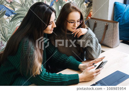 Gen Z influence on the development and popularity of short-form video content on social media platforms. Two girlfriend in cozy sweaters share a moment over smartphone and record video 108985954