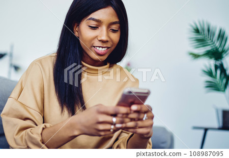 Smiling millennial african american woman sitting on sofa in living room with cellular connected to wifi Smiling millennial african american woman sitting on sofa in living room with cellular connected to wifi 108987095