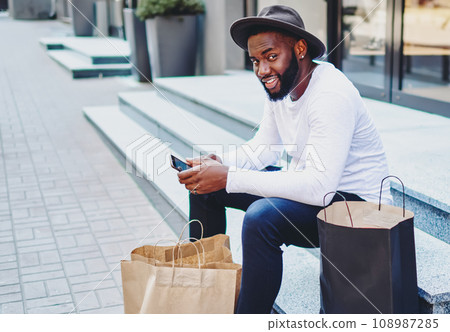 positive dark skinned man in stylish hat resting after shopping time positive dark skinned man in stylish hat resting after shopping time 108987285