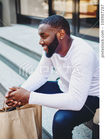 Smiling afro american male customer in trendy wear sitting on stairs of store with bags Smiling afro american male customer in trendy wear sitting on stairs of store with bags 108987287