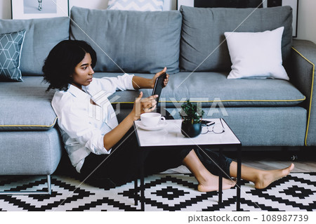 African american young woman dressed in casual wear sitting on floor in modern flat with stylish interior 108987739