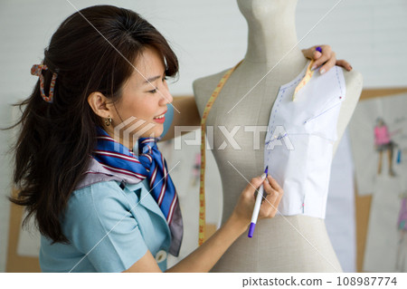 An Asian woman is carefully sewing clothes on a manufactured figure, known as a mannequin. An Asian woman is carefully sewing clothes on a manufactured figure, known as a mannequin. 108987774