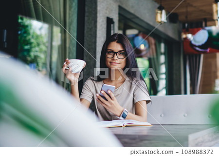 hipster girl looking at camera while chatting online on smartphone hipster girl looking at camera while chatting online on smartphone 108988372