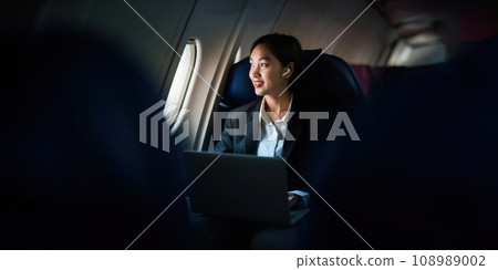 Successful Asian business woman, Business woman working in airplane cabin during flight on laptop computer listening to music with headphones 108989002