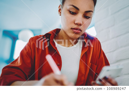 serious hipster girl doing school homework while updating smartphone gadget serious hipster girl doing school homework while updating smartphone gadget 108989247