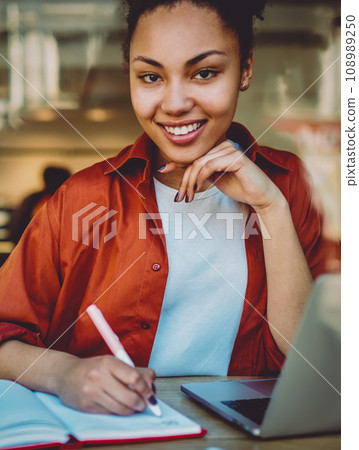 portrait of smiling african woman planning month in notepad and using organiser on netbook 108989250