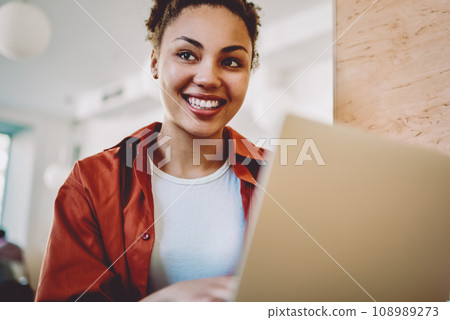 positive black woman looking away and smiling during e learning in cafe interior positive black woman looking away and smiling during e learning in cafe interior 108989273