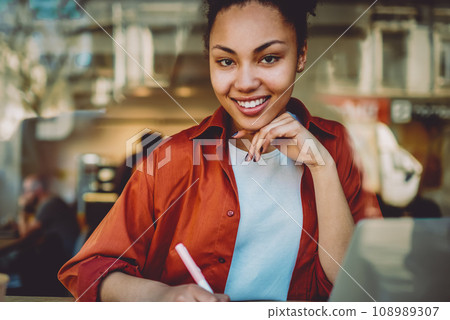 portrait of smiling african woman planning month in notepad and using organiser on netbook 108989307
