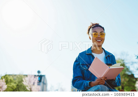 Positive female student in sunglasses enjoying studying on nature with textbook for education 108989415