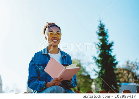 Positive female student in sunglasses enjoying studying on nature with textbook for education Positive female student in sunglasses enjoying studying on nature with textbook for education 108989417