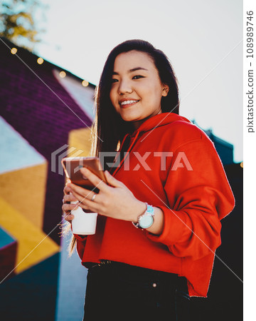 Positive young woman dressed in casual wear installing app on mobile phone while smiling at camera Positive young woman dressed in casual wear installing app on mobile phone while smiling at camera 108989746