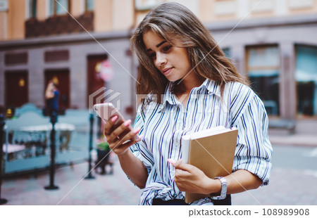 Female student reading message from modern mobile phone strolling on street Female student reading message from modern mobile phone strolling on street 108989908