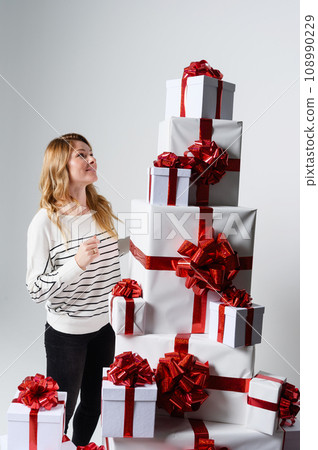 Beautiful woman poses among a mountain of white gift boxes in a gray background. Beautiful woman poses among a mountain of white gift boxes in a gray background. 108990229