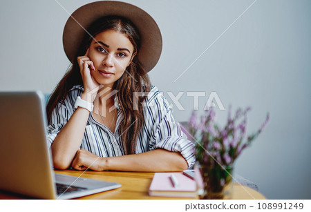 beautiful pensive hipster girl in hat looking at camera working on freelance via netbook 108991249