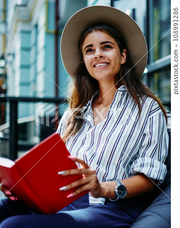 cheerful hipster girl in stylish hat spending free time on hobby holding interesting best seller 108991286