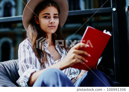 Serious female in trendy hat looking at camera sitting with book having free time to spent on hobby Serious female in trendy hat looking at camera sitting with book having free time to spent on hobby 108991306