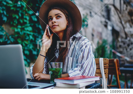 Pensive hipster girl in trendy hat sitting at table on street cafe talking on mobile phone 108991350