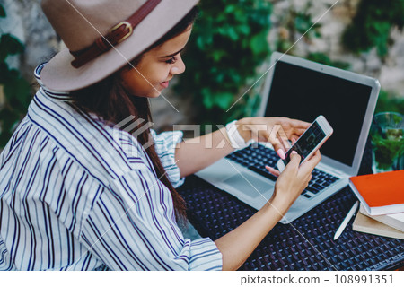 Positive hipster girl in hat checking new feed in social networks using mobile phone and 4G connection Positive hipster girl in hat checking new feed in social networks using mobile phone and 4G connection 108991351
