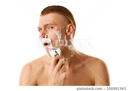 Facial grooming. Serious male model, attractive guy shaving his beard with razor and shaving cream against white background. Facial grooming. Serious male model, attractive guy shaving his beard with razor and shaving cream against white background. 108991363