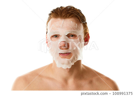 Procedures for glowing skin. Red haired young man with moisturizer, hydrating mask on face against white studio background. 108991375
