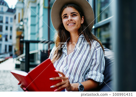cheerful hipster girl in stylish hat spending free time on hobby holding interesting best seller cheerful hipster girl in stylish hat spending free time on hobby holding interesting best seller 108991431