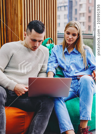 Young man and woman dressed in casual wear sitting on comfortable poufs watching film online 108991936