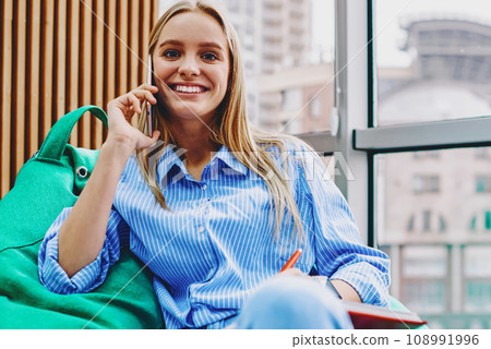 Cheerful hipster blogger laughing during phone conversation on cellular sitting on pouf Cheerful hipster blogger laughing during phone conversation on cellular sitting on pouf 108991996