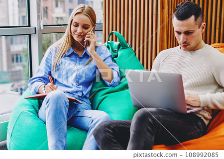 Hipster girl calling on smartphone and noting information in notebook sitting on pouf while fer boyfriend  108992085