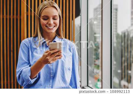 Positive young woman chatting online on cellular standing in office interior Positive young woman chatting online on cellular standing in office interior 108992113