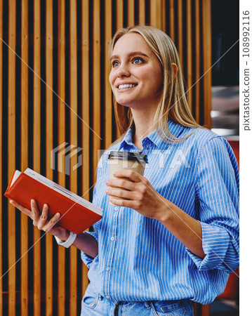 Dreamy young woman with blonde hair holding opened book and tasty coffee beverage in hands Dreamy young woman with blonde hair holding opened book and tasty coffee beverage in hands 108992116